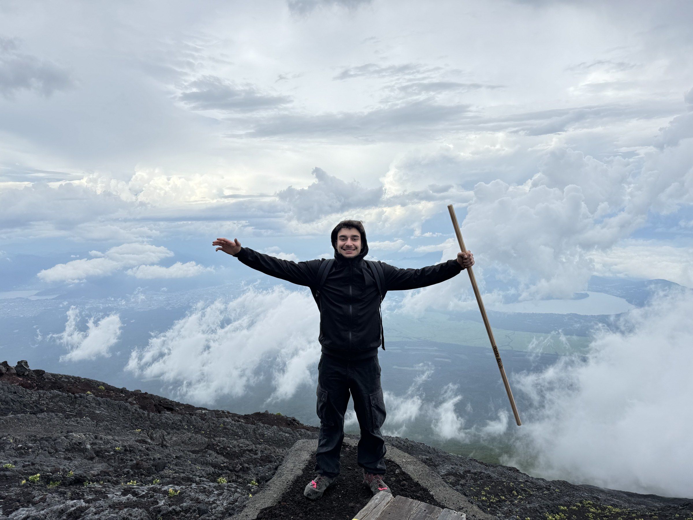 Orhun with arms wide on the slopes of Mount Fuji above the clouds, holding a walking stick