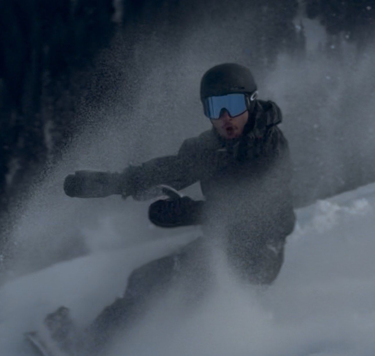 Orhun snowboarding through a spray of powder, goggles on, close to the camera